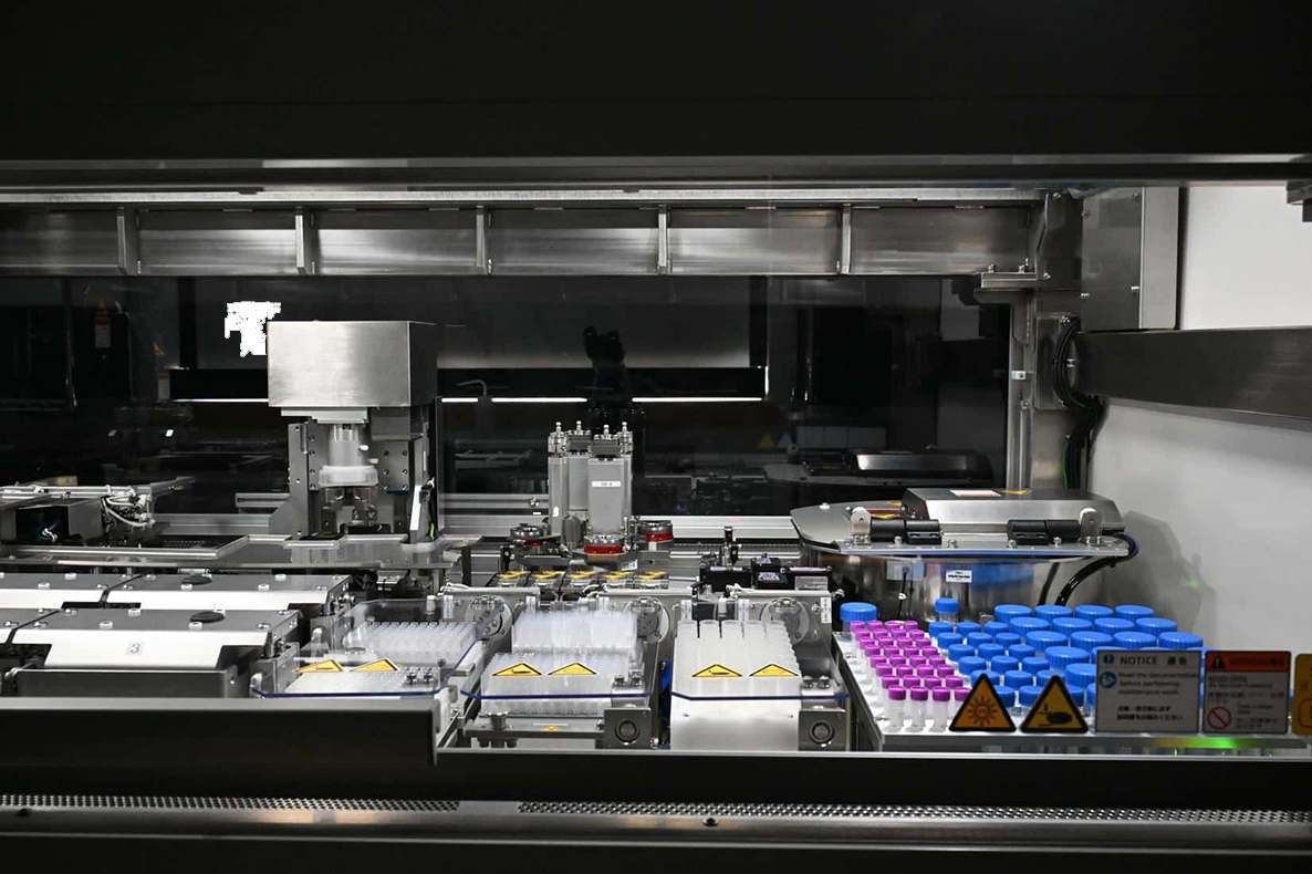 Interior of the NPi-11 cell culture robot showing robotic liquid handling arms, blue centrifuge tube racks, and plate handling area during automated cell culture operation.
