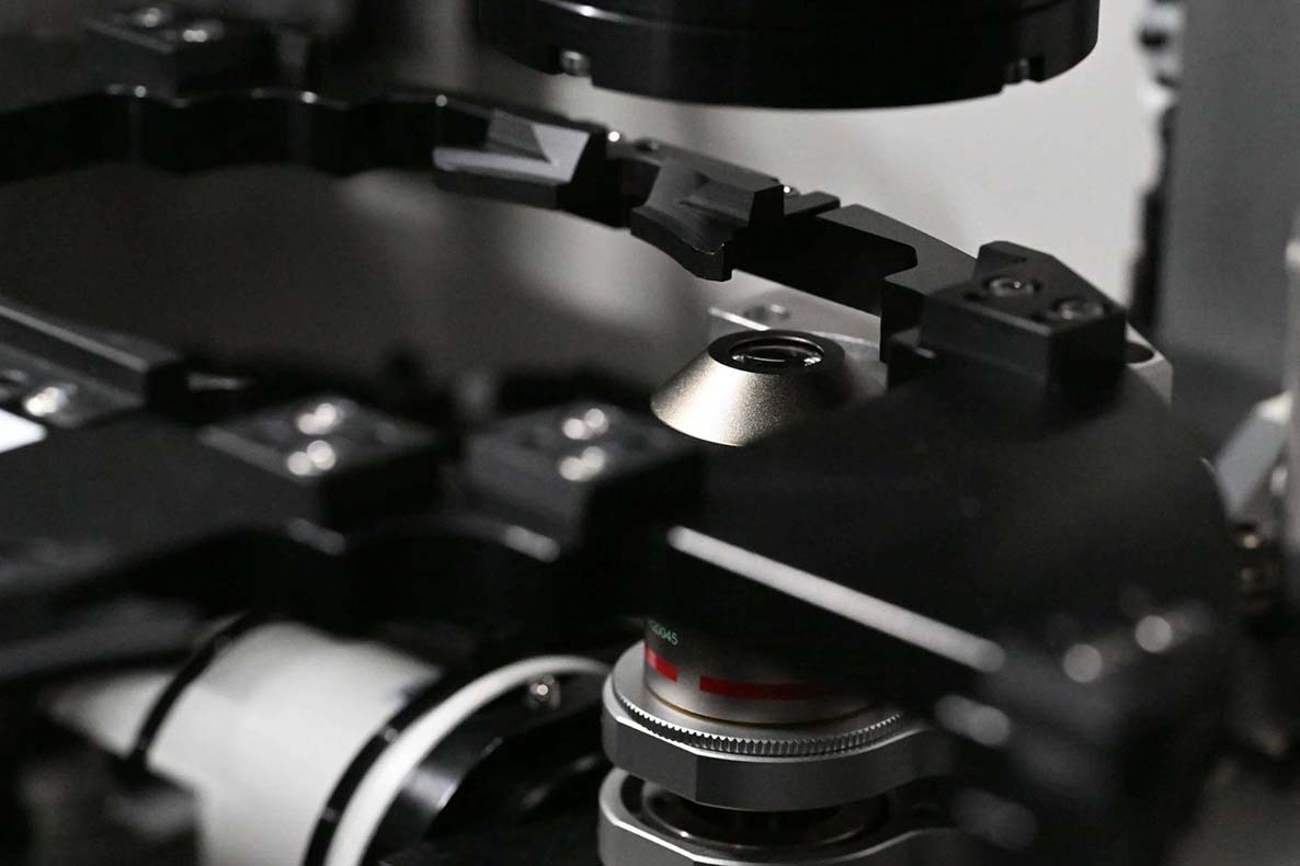 Close-up of the NPi-11 cell culture robot's built-in phase-contrast microscope objective lens positioned over the imaging stage for automated cell analysis.