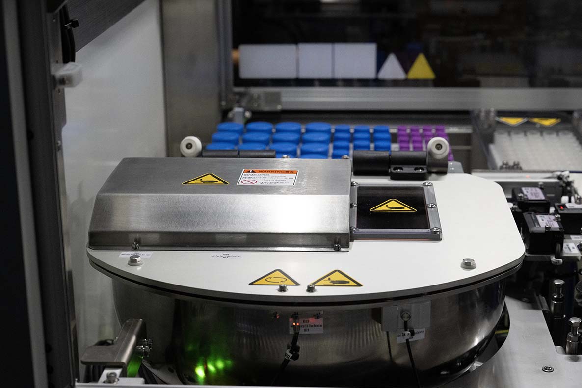 Close-up of the NPi-11 automated cell culture system's built-in centrifuge with stainless steel housing and safety labels, with racks of blue-capped and pink-capped centrifuge tubes visible in the background.