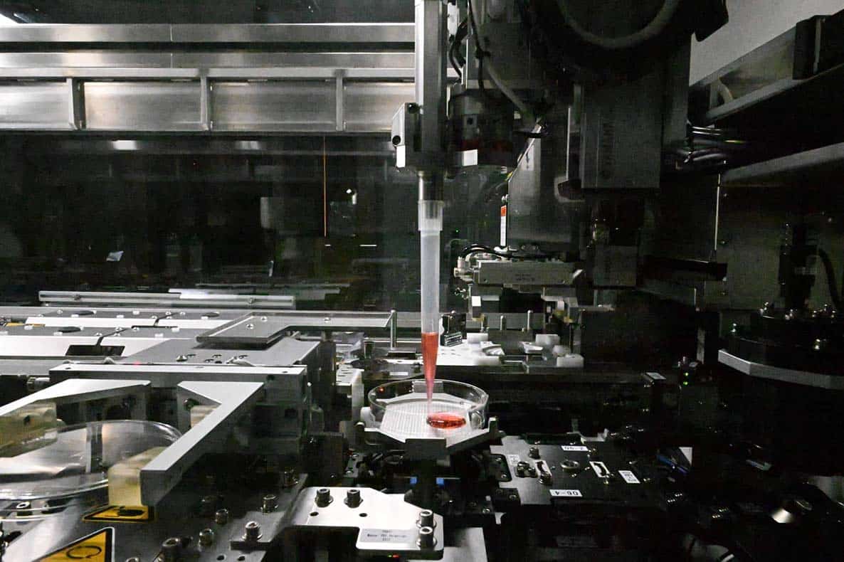 Close-up of the NPi-11 cell culture robot's robotic pipetting arm dispensing pink cell culture media from a serological pipette into a cell culture dish inside the automated cell culture system's enclosed workspace.